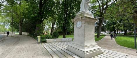 Une statue dans un parc, surmontée d’une horloge, entourée d’arbres et bordée d’une allée pavée.