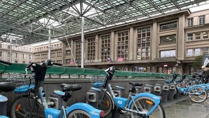 Uma área de estacionamento coberta para bicicletas, com várias bicicletas azuis, um edifício grande com janelas amplas ao fundo e uma placa com a inscrição 'SAFETY OF AMIENS'.