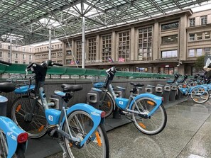A covered bike parking area with multiple blue bicycles, a large building with large windows in the background, and a sign that reads 'SAFETY OF AMIENS.' - Queen Bed • Fully Equipped Apartment • Washing Machine • Amiens Centre • 5-min Walk to Station • Parking on Site (Amiens)