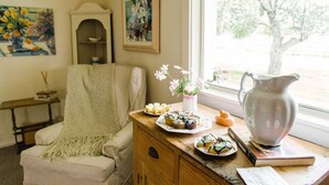 Ein gemütliches Zimmer mit Stuhl, Tisch mit Speisen, Blumen in einer Vase und einem Fenster mit Blick auf Bäume.