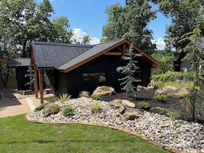 A modern cabin with a sloped roof, surrounded by a landscaped garden with rocks and plants. - Lakefront Escape in Hot Springs 2BR/2BA, Hot Tub, Fireplace & Boat Slips (Hot Springs National Park)