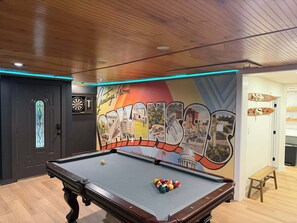 A pool table with colorful balls, a dartboard, and a mural with the word 'Greetings' on the wall.