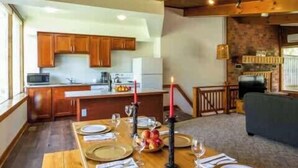 Un coin repas avec une longue table en bois dressée pour quatre personnes, une cuisine en arrière-plan et un espace de vie chaleureux agrémenté d’une cheminée.