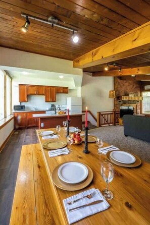 A long wooden dining table set for a meal, a kitchen area with wooden cabinets, and a stone fireplace.
