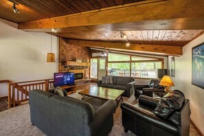 A cozy living room with a fireplace, leather sofas, a flat-screen TV, and a large window with a view of trees.