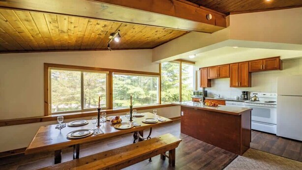 Une table à manger en bois avec des bancs, un coin cuisine doté de meubles en bois et une grande fenêtre offrant une vue sur les arbres.