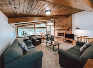A cozy living room with a fireplace, wooden ceiling, and large windows.