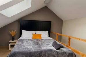 Une chambre avec un plafond en pente, un lit doté d’une tête de lit sombre, des coussins blancs et orange, une couverture grise, une table de chevet en bois avec un vase, et un rebord de fenêtre en bois.