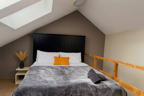 A bedroom with a sloped ceiling, a bed with a dark headboard, white and orange pillows, a gray blanket, a wooden nightstand with a vase, and a wooden railing on the window.