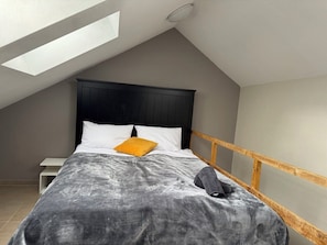Une chambre avec un plafond en pente, un lit recouvert d’une couverture grise, une tête de lit en bois et une petite table de chevet.