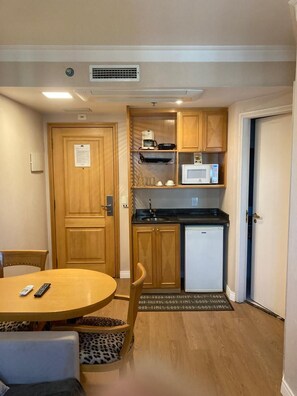 A compact kitchen with wooden cabinets, a microwave, and a refrigerator. There is a dining table with chairs and a remote control on it. - Lume Pinheiros Flat (São Paulo)