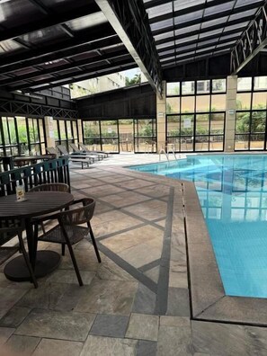An indoor swimming pool with a tiled floor, surrounded by lounge chairs and a covered seating area. - Lume Pinheiros Flat (São Paulo)