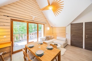 Une pièce avec un plafond en bois, une table à manger dressée pour quatre personnes, un canapé et une grande fenêtre donnant sur un paysage verdoyant.