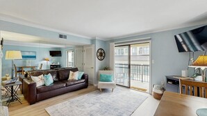 Ein Wohnzimmer mit einem braunen Ledersofa, einem weißen Sessel, einem Esstisch, einem Flachbildfernseher und einem großen Fenster mit Blick auf Gebäude.