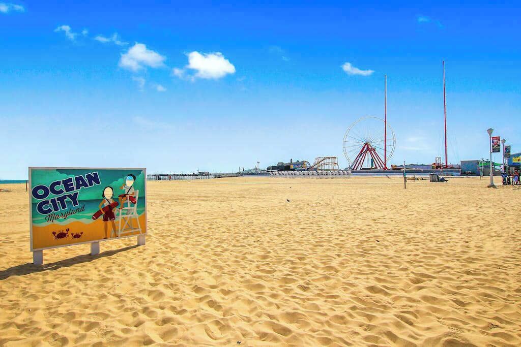 A beach with a sign reading 'Ocean City Maryland,' a Ferris wheel, and a boardwalk in the background.