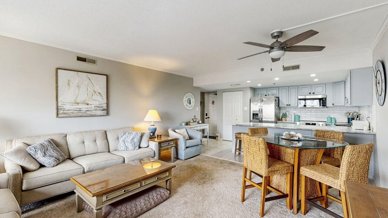 A living room with a sofa, coffee table, and dining area with chairs. There is a kitchen in the background with cabinets and appliances.
