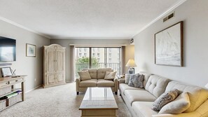A living room with a sofa, a coffee table, a cabinet, a TV, and a window with a view of buildings. - Royal Hawaiian 205 (Ocean City)