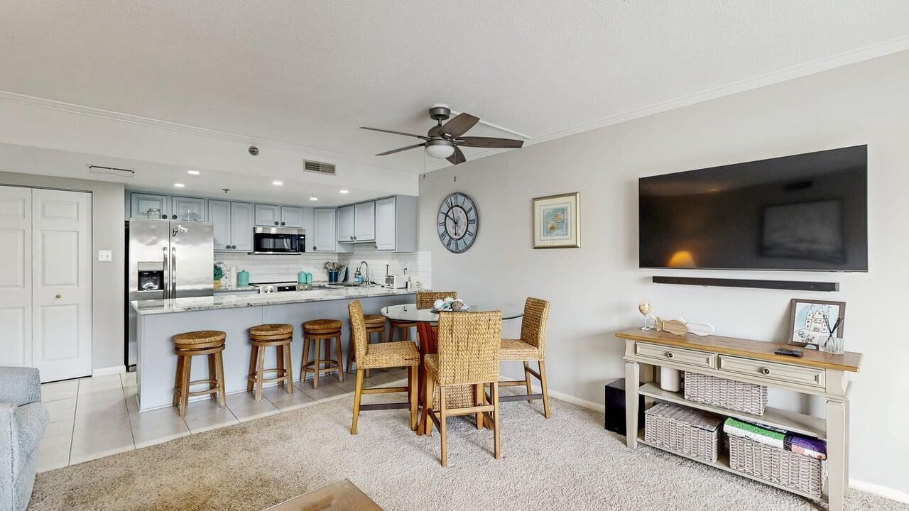 A modern kitchen with a central island, bar stools, a dining table with chairs, a ceiling fan, a mounted TV, and a clock on the wall.