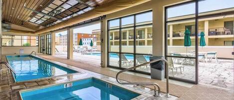 Piscina interna com paredes de vidro transparente, teto de madeira e vista para edifícios externos.