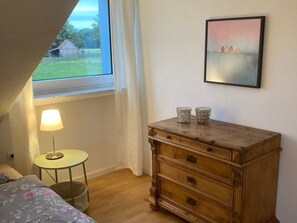 A bedroom with a wooden dresser, a lamp, a framed picture, and a window with a view of a barn and trees. - Leling Holiday Apartment – Relaxation with a Wellness View (Havixbeck)
