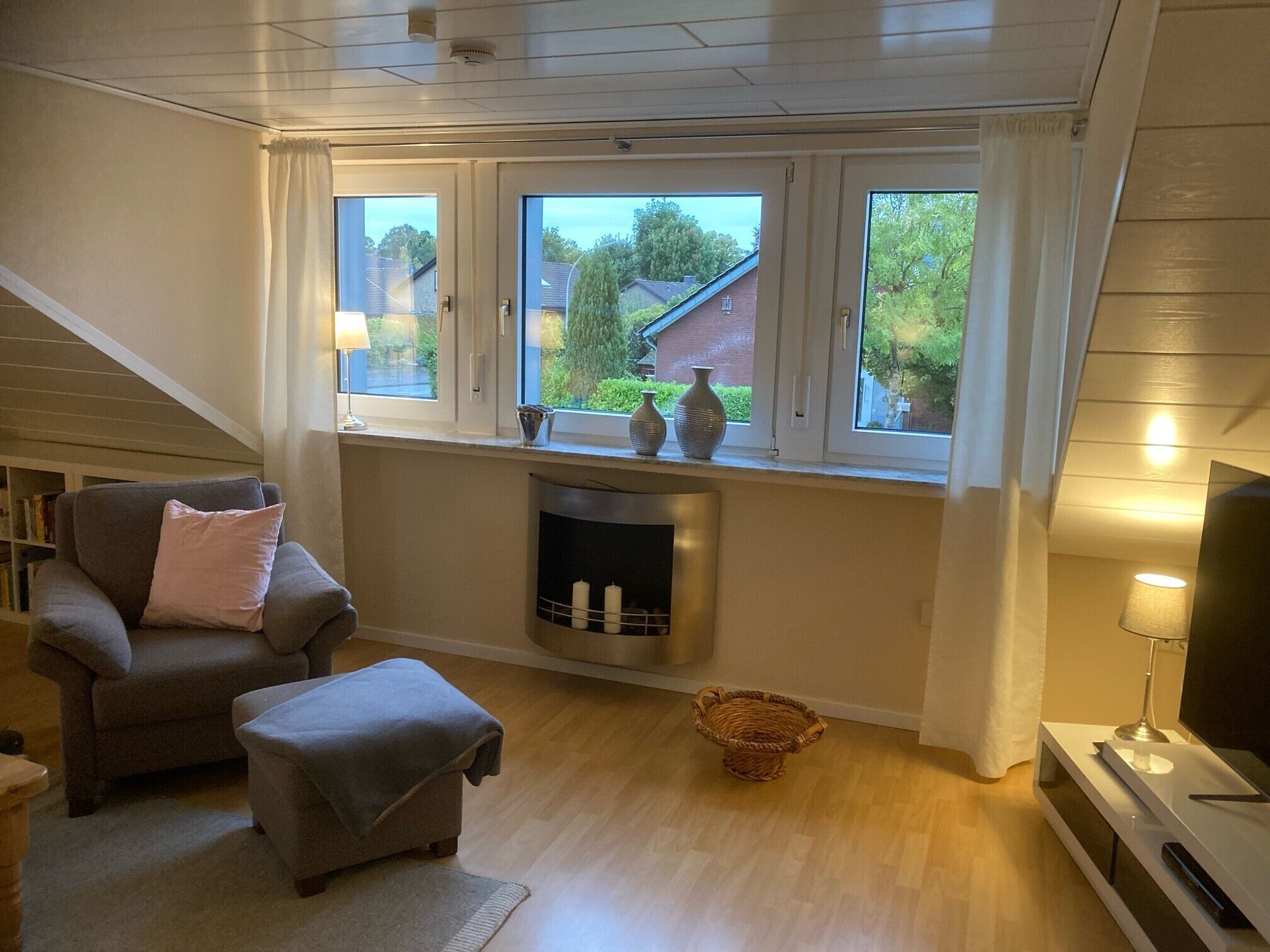 Ett vardagsrum med en öppen spis, en soffa, en tv och ett fönster med utsikt över ett hus och träd.