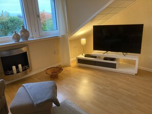 A modern living room with a fireplace, a television on a white stand, and a view of the outdoors through a large window. - Leling Holiday Apartment – Relaxation with a Wellness View (Havixbeck)