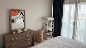 A bedroom with a bed, a dresser, a mirror, a shelf with towels, a round stool, and a window with curtains. - Modern Comforts: Al Raha Beach modern 1 bedroom near AD airport (Abu Dhabi)