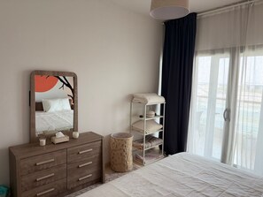 Ein Schlafzimmer mit einem Bett, einer Kommode, einem Spiegel, einem Handtuchregal, einem runden Hocker und einem Fenster mit Vorhängen.
