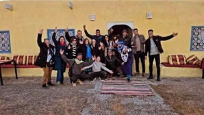 Un grupo de personas reunidas en un espacio al aire libre con un muro amarillo, una puerta decorada y un adorno mural con motivos.