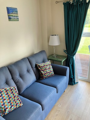 Ein blaues Sofa mit bunten Kissen, ein kleiner Tisch mit einer Lampe und ein Blick ins Grüne durch ein Fenster mit Vorhängen.