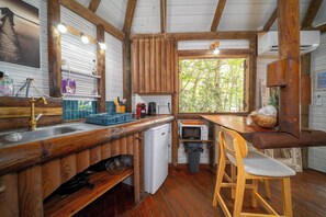 A rustic kitchen with wooden beams, a sink, a microwave, and a window with a view of trees. - Bungalow Yuka, a refuge between sea and nature (Les Anses-d'Arlet)