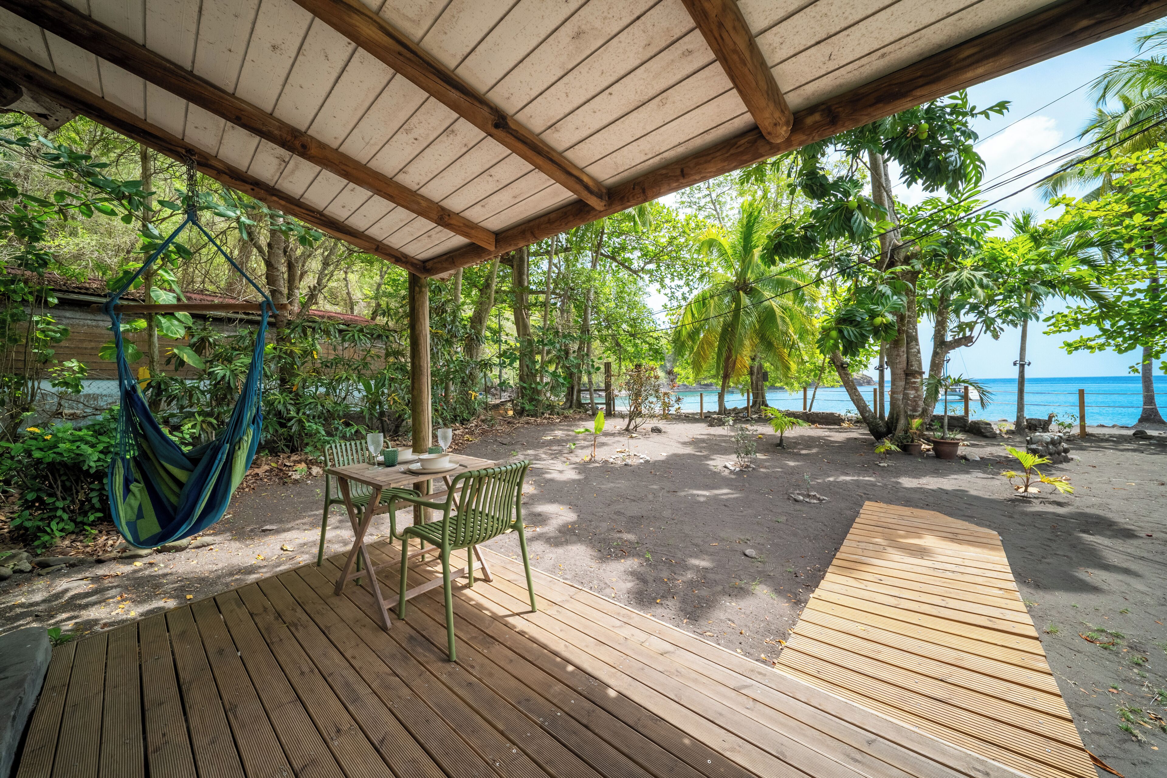 A wooden deck with a hammock, a table, and chairs, overlooking a beach with palm trees and the ocean.