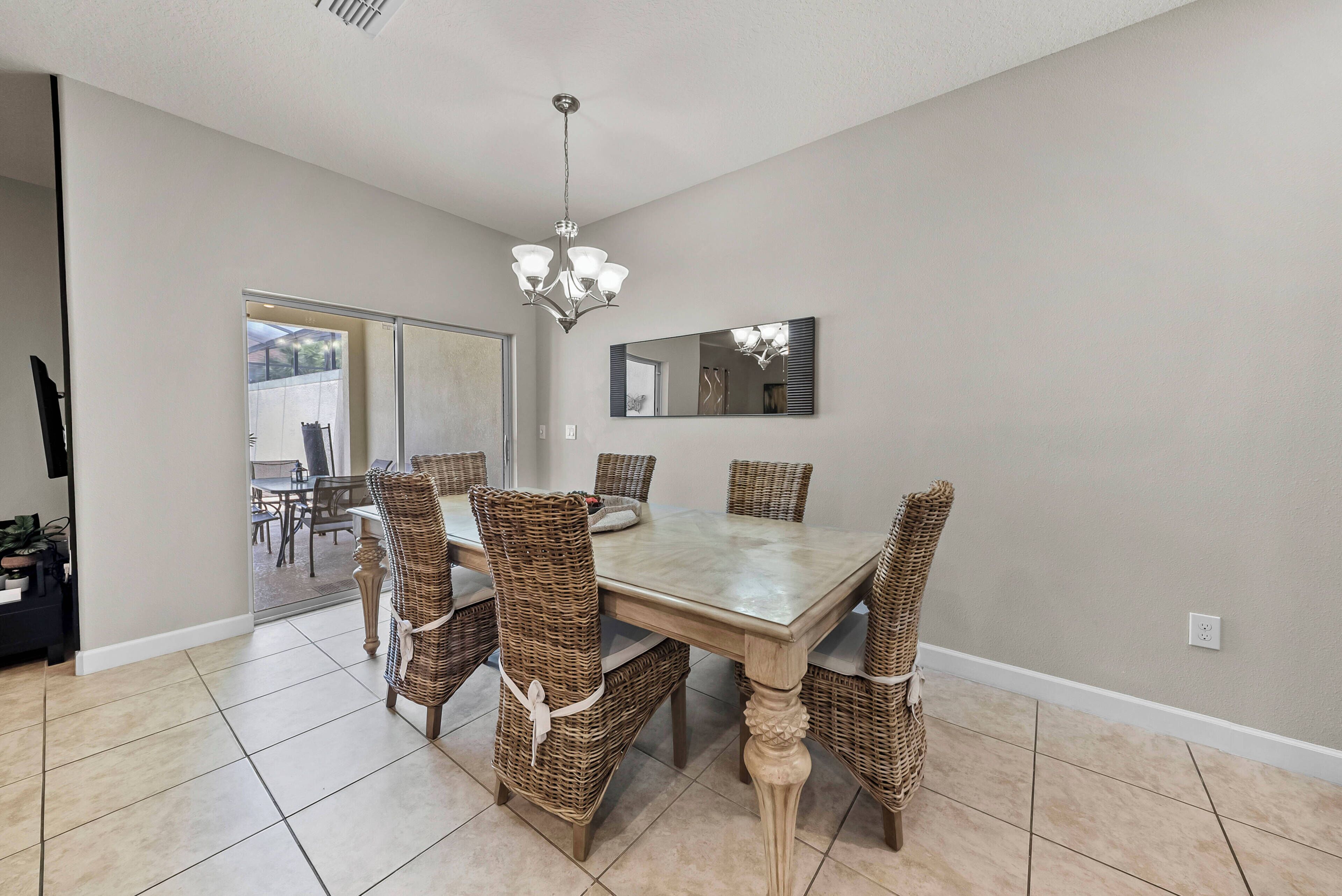 A dining area with a large table and wicker chairs, a chandelier, and a sliding glass door leading to an outdoor patio.