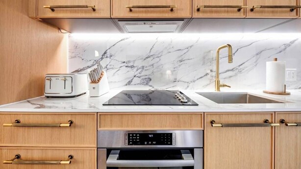 Una cocina con armarios de madera, salpicadero de mármol, cocina de inducción negra, horno de acero inoxidable y grifo dorado.