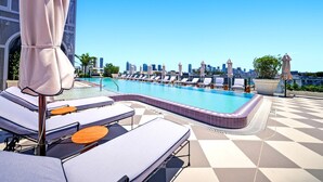Una piscina en la azotea con sillones de descanso y sombrillas, con un horizonte urbano al fondo y un cielo azul despejado.
