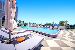 Ein Pool auf dem Dach mit Liegestühlen und Sonnenschirmen, im Hintergrund ein Stadtbild und ein klarer, blauer Himmel.