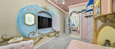 A bedroom with a large mirror, a television, a plush pink chair, a bed with a pink bedspread, and a castle-themed play area.