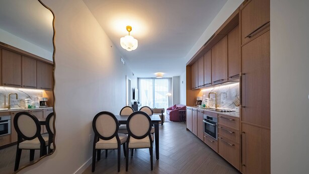 A modern kitchen with wooden cabinets, a dining area with chairs, and a large window providing natural light. - Apartment at Nomad Residences Wynwood with 2BR (Miami)