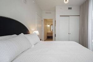 Ein Hotelzimmer mit einem großen Bett, einer Nachttischlampe, einer Tür zu einem Badezimmer und einem Fenster mit Vorhängen.