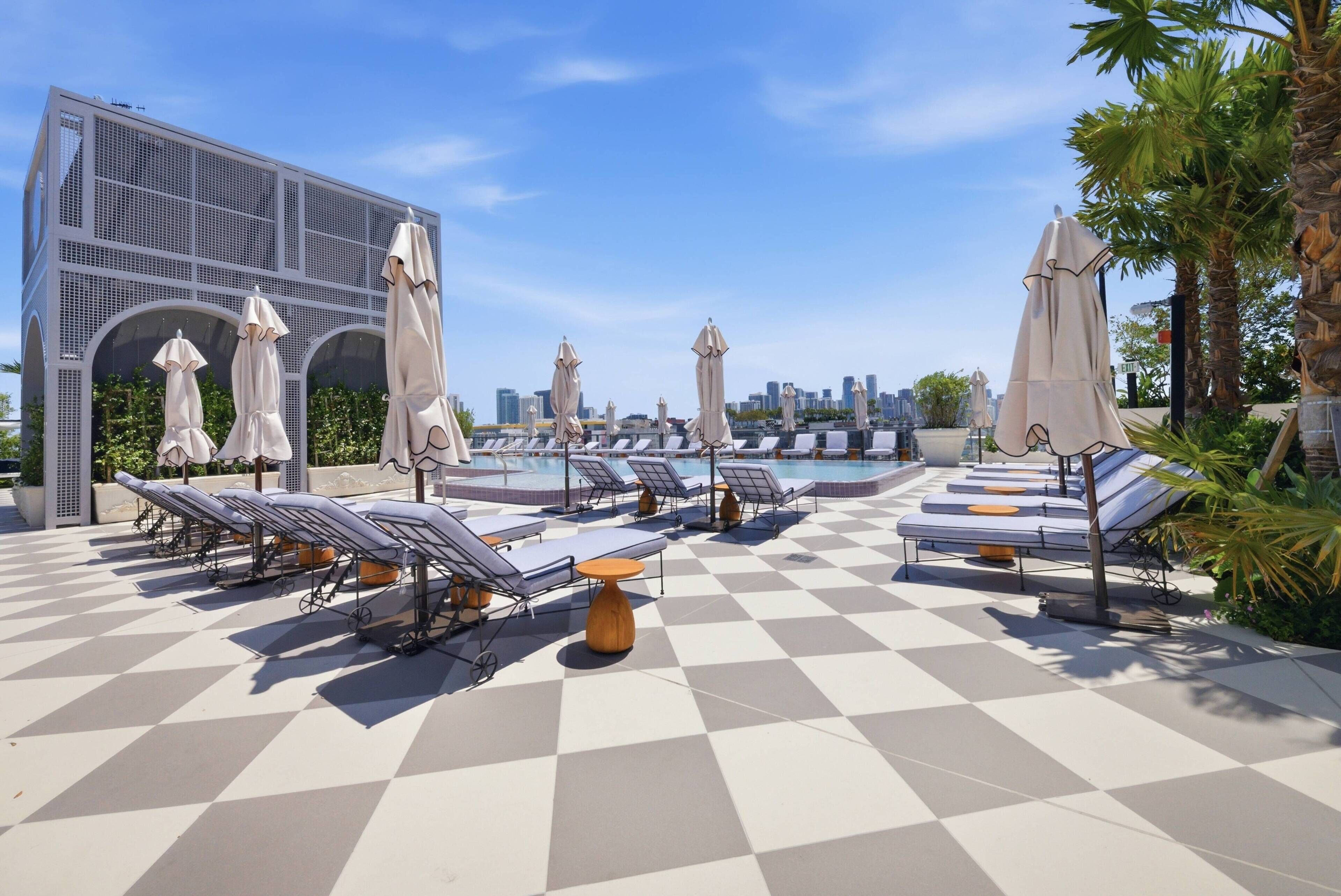 A rooftop pool area with sun loungers and umbrellas, a checkered floor, and a city skyline in the background.