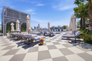 Ein Poolbereich auf dem Dach mit Liegestühlen und Sonnenschirmen, einem Schachbrettmuster auf dem Boden und einer Stadtansicht im Hintergrund.