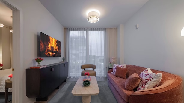 En moderne stue med sofa, sofabord, fjernsyn ophængt på væggen og et stort vindue med lette gardiner.