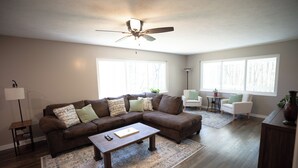 A living room with a sectional sofa, a coffee table, a floor lamp, a ceiling fan, and large windows. - Secluded 5BR Retreat w/ Hot Tub, Sleeps 10 (Canfield)
