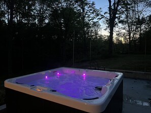 Una vasca idromassaggio illuminata di viola, circondata da alberi e un cielo sereno.