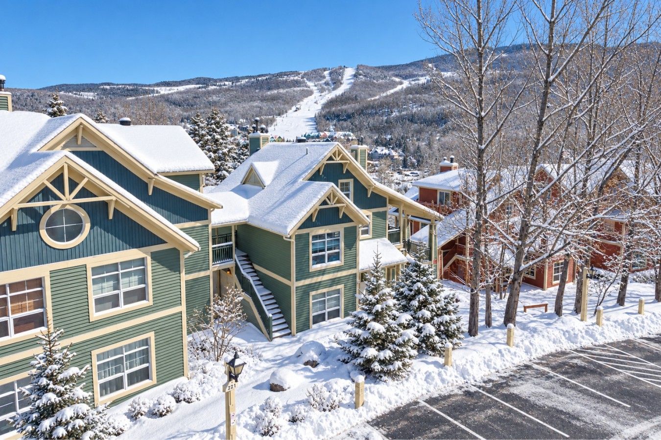 A snow-covered residential area with houses, a parking lot, and a ski slope in the background.