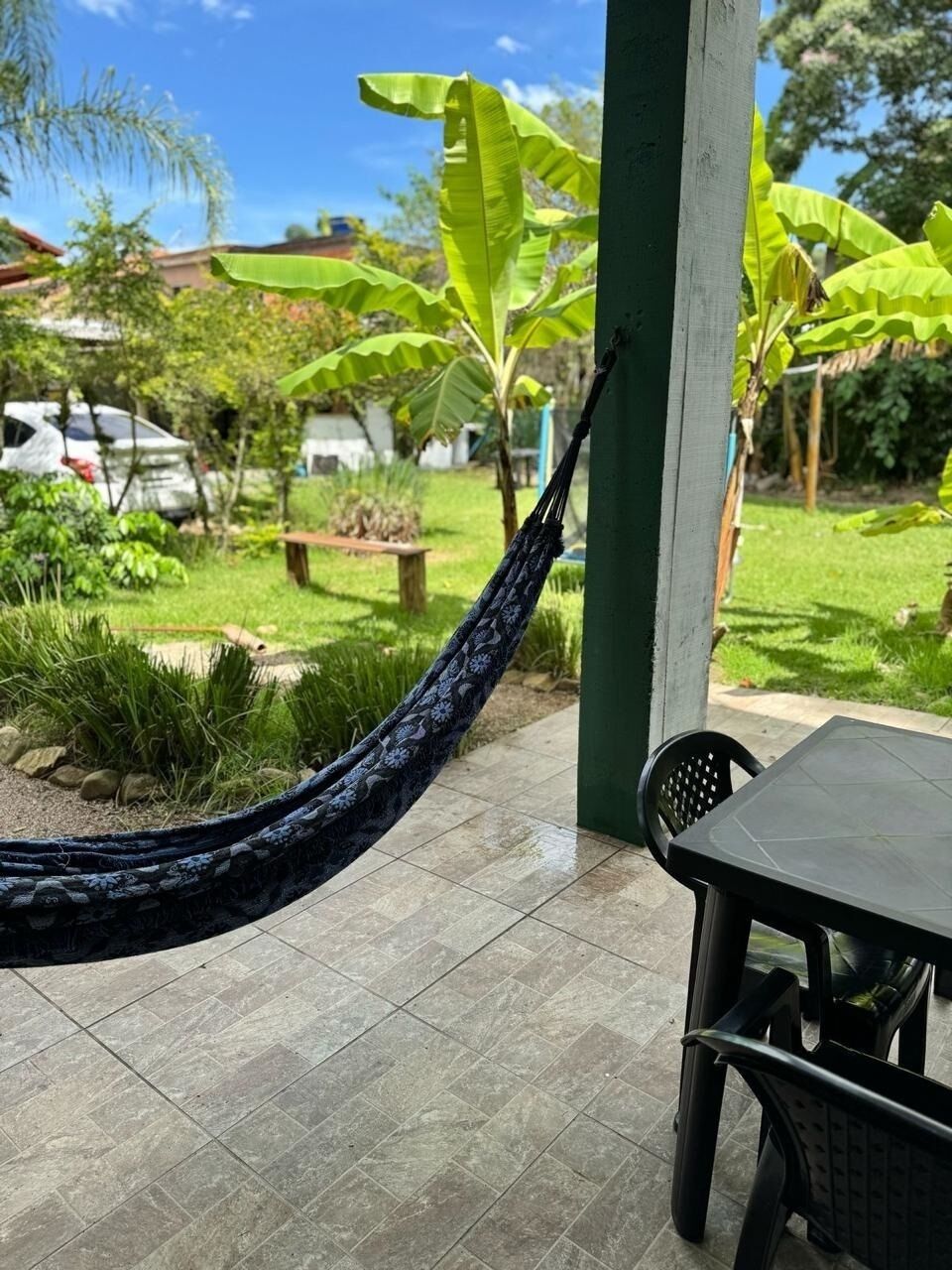 A hammock hanging over a tiled patio with a table and chairs, surrounded by lush greenery and banana trees.