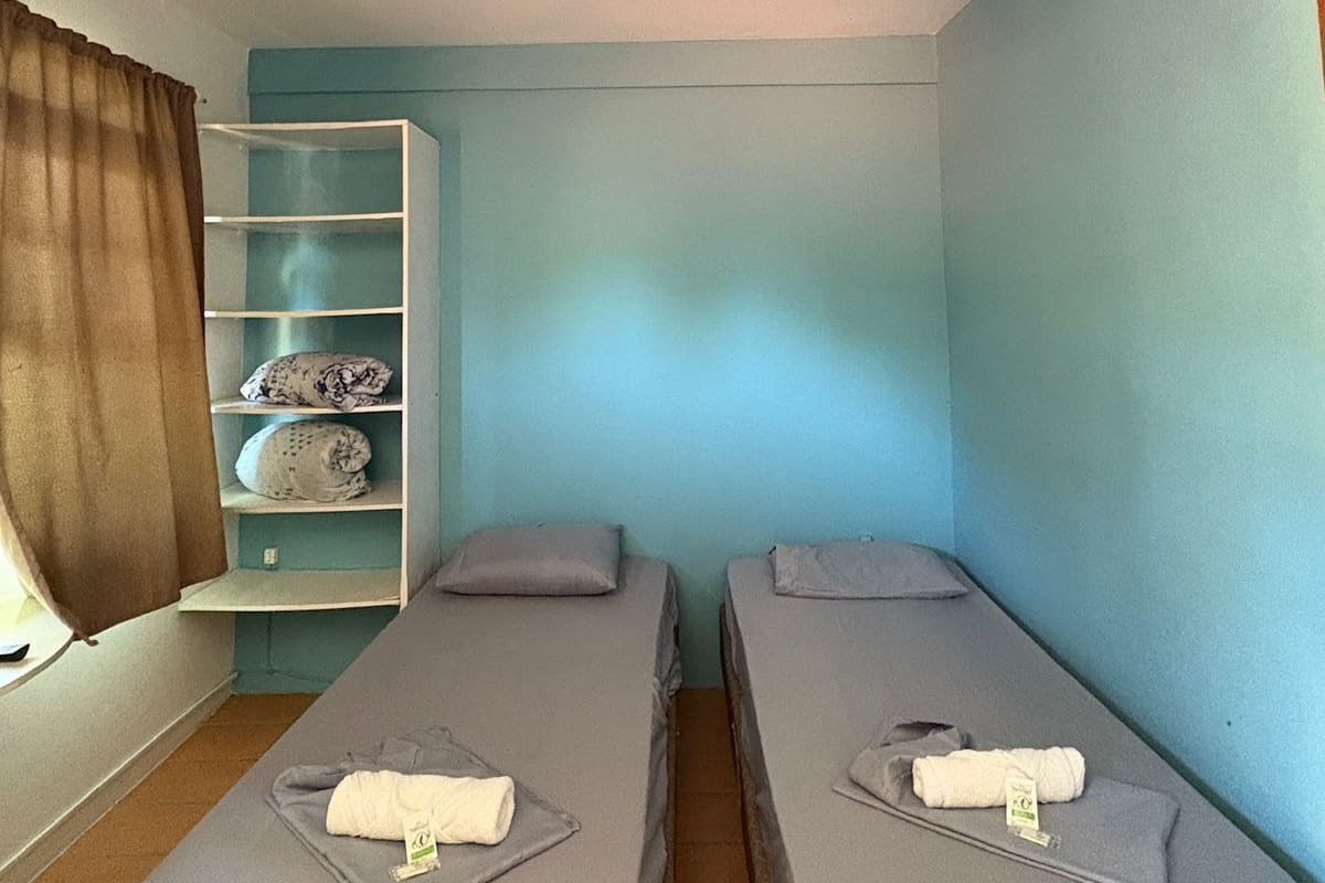 Una habitación pequeña con dos camas, un ventilador de techo, un estante con objetos y una ventana con cortinas.