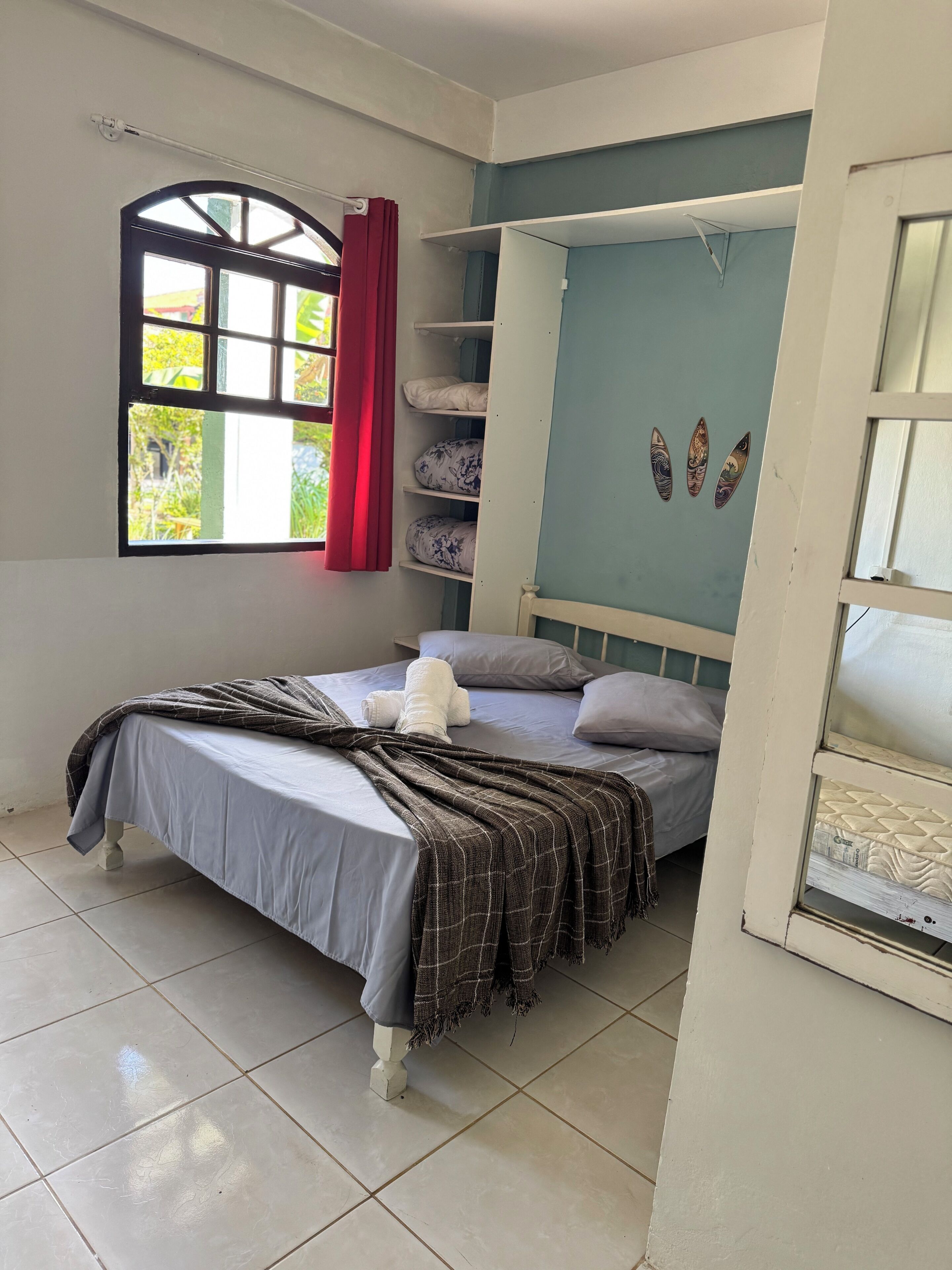 Une chambre à coucher avec un lit, une fenêtre avec des rideaux, une étagère avec des coussins et un mur décoré de différents objets.