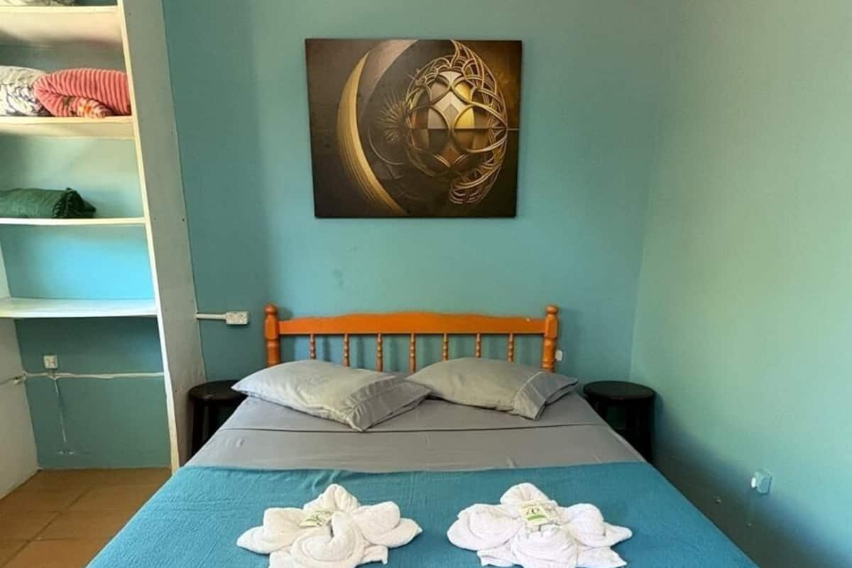 Un dormitorio con una cama, un edredón azul, almohadas, una obra de arte en la pared y una estantería con ropa doblada.