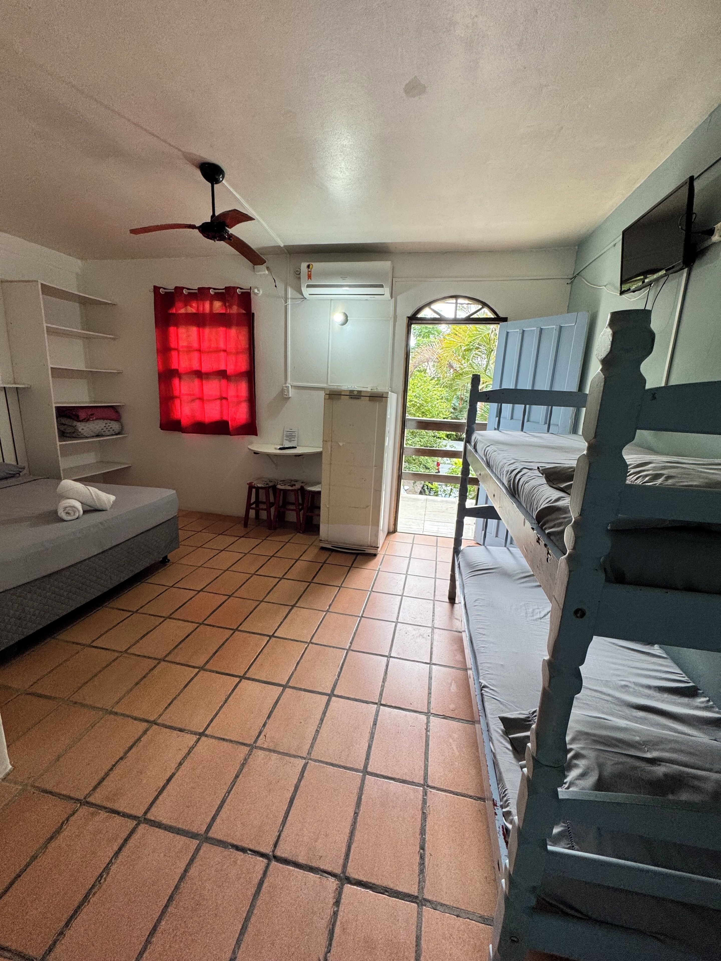 A dormitory room with a bed, a desk, a chair, a shelf, a ceiling fan, and a window.